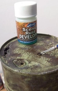 Flacon Scenic Rust Developer aplicat cu pensula pe o machetă de butoi metalic, efect de patinare și coroziune