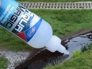 Aplicarea Aqua Magic direct din flacon pe o machetă feroviară, creând efect de apă într-un șanț lângă calea ferată