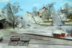 Machetă feroviară cu peisaj de sat și efect de zăpadă subțire pe copaci, acoperișuri și teren, creat cu Scenic Snow Kit