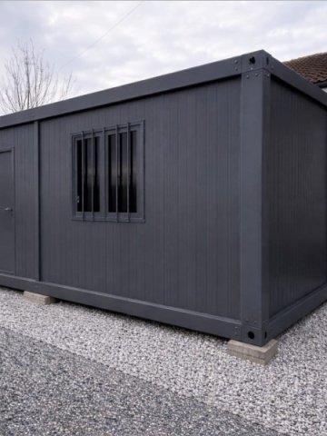 Container antracit cu ușă plină și fereastră cu bare, amplasat pe pietriș