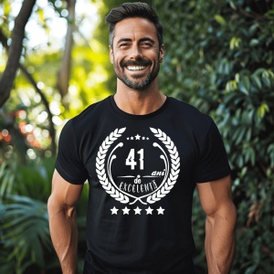 Un bărbat zâmbitor purtând un tricou negru cu textul ''41 de ani de excelență'', înconjurat de o coroană de lauri.