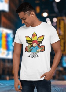 Bărbat zâmbitor purtând un tricou alb cu un design vibrant ce include un personaj urban stilizat cu spray-uri de graffiti și un logo reinterpretat în galben-roșu, pozând într-un cadru urban luminat cu reclame și lumini de noapte.