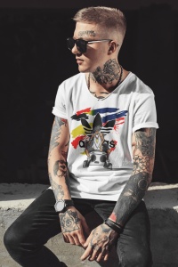 Bărbat tatuat, purtând ochelari de soare și un tricou alb cu un design urban, ce include un personaj cu un boombox și un fundal colorat abstract, pozând într-un cadru minimalist, luminat natural.