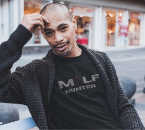Un bărbat relaxat poartă un tricou negru cu textul 'MILF HUNTER' în font gri și o ilustrație stilizată în roșu, alături de un cardigan gros. Fundalul este un spațiu urban, cu vitrine de magazine în spate.
