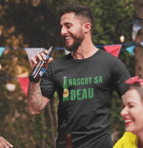 Bărbat zâmbitor, purtând un tricou negru cu mesajul 'Născut să Beau' și imaginea unei sticle de bere. Ține o sticlă în mână și pare să se bucure de o petrecere în aer liber, decorată cu ghirlande colorate și lumini în fundal.