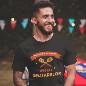 Bărbat zâmbitor purtând un tricou negru cu mesajul 'Familia mea mă numește Regele Grătarelor,' decorat cu grafică de ustensile de grătar. În fundal, sunt vizibile decorațiuni colorate de petrecere, creând o atmosferă festivă în aer liber.