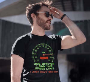 Un bărbat purtând ochelari de soare poartă un tricou negru cu un design ce afișează un vitezometru și mesajul amuzant 'Yes officer, I saw the speed limit, I just didn't see you' în text verde și roșu. Bărbatul își ridică mâna spre frunte, iar fundalul est