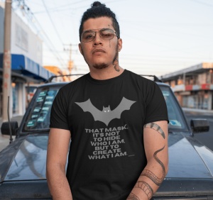 Bărbat cu ochelari de vedere și tatuaje pe gât și braț, purtând un tricou negru cu un design inspirat de Batman, reprezentând o siluetă de liliac și textul: 'THAT MASK. IT'S NOT TO HIDE WHO I AM, BUT TO CREATE WHAT I AM.' În spate se află o mașină și un p
