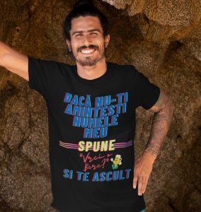 Un bărbat zâmbitor poartă un tricou negru cu mesajul amuzant 'Dacă nu-ți amintești numele meu, spune „Vrei bere?” și te ascult', colorat în albastru, galben și roșu. Bărbatul are părul prins și stă sprijinit pe o suprafață de stâncă.