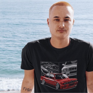 Bărbat purtând un tricou negru cu o ilustrație grafică detaliată a unui BMW roșu clasic, pozat pe fundalul unei plaje cu oceanul în fundal.