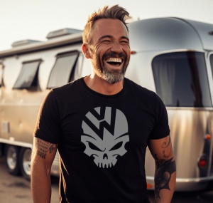 Bărbat matur zâmbitor, cu barbă și tatuaje pe brațe, purtând un tricou negru cu design de craniu stilizat și sigla Volkswagen, pozând lângă o rulotă modernă reflectorizantă sub lumina soarelui.