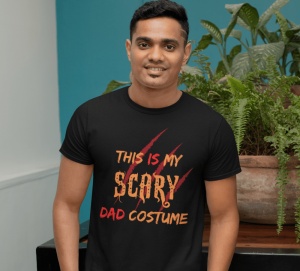 Un bărbat care zâmbește, purtând un tricou negru cu textul 'This is my scary dad costume' în nuanțe de portocaliu și roșu, completat cu un design de zgârieturi de gheare. Bărbatul stă în fața unor plante verzi într-un decor interior.