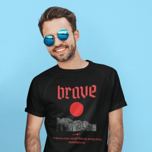 Un bărbat zâmbitor poartă ochelari de soare rotunzi și un tricou negru cu inscripția 'brave' în roșu, însoțită de o imagine grafică cu un soare și un peisaj montan stilizat. Fundalul este un perete albastru vibrant, adăugând contrast și evidențiind design