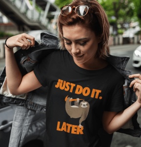 Tânără purtând un tricou negru cu un design care înfățișează un leneș atârnând de o ramură și textul 'JUST DO IT. LATER' scris în portocaliu. Ea poartă o jachetă de blugi descheiată, care este trasă pe umeri, și privește în jos cu un zâmbet subtil. Fundal
