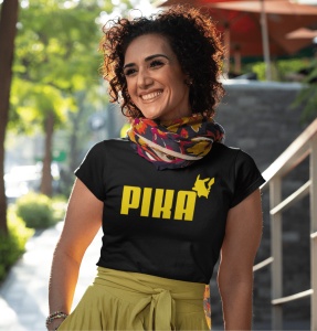 Femeie zâmbitoare purtând un tricou negru cu design galben ce include textul 'PIKA' și o siluetă stilizată a unui personaj în colțul din dreapta al textului. Ea poartă un fular colorat și o fustă galbenă, iar fundalul arată o stradă cu verdeață, creând o 