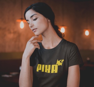 Tânără purtând un tricou negru cu un design galben ce include textul 'PIKA' și o siluetă stilizată a unui personaj în colțul din dreapta al textului. Ea își sprijină mâna pe bărbie și pare să fie adâncită în gânduri, privind ușor în jos. Fundalul este dec