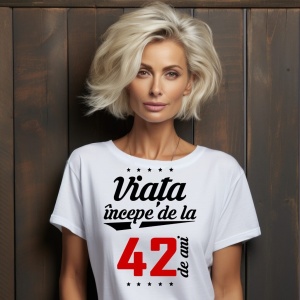 O femeie purtând un tricou alb cu textul  'Viața începe de la 42 de ani'. Fundal din lemn cu diferite nuanțe de maro și verde.