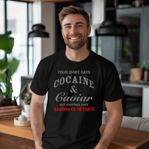 Bărbat zâmbitor, cu barbă scurtă, purtând un tricou negru cu text amuzant: 'YOUR SHIRT SAYS COCAINE & CAVIAR BUT YOUR FACE SAYS ȘAORMA CU DE TOATE', stând într-un interior modern decorat cu plante și mobilă elegantă.