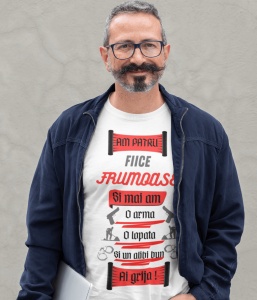 Bărbat zâmbitor purtând un tricou alb cu mesajul 'Am patru fiice frumoase și mai am o armă, o lopată și un alibi bun. Ai grijă!', însoțit de ilustrații de arme, lopată și cătușe. Bărbatul poartă o geacă bleumarin deschisă și ochelari, stând în fața unui f