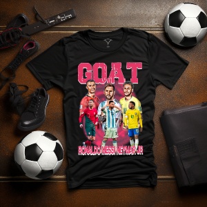 Tricou negru cu designul text 'GOAT' și portrete ale unor jucători celebri de fotbal, așezat pe o suprafață alături de mingi de fotbal, ghete și accesorii sportive.