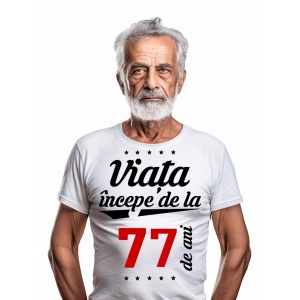 Bărbat în vârstă cu barbă albă, purtând un tricou alb cu mesajul 'Viața începe de la 77 de ani', pe un fundal alb simplu.