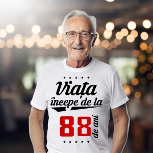 Bărbat în vârstă purtând ochelari și un tricou alb cu mesajul 'Viața începe de la 88 de ani', într-un cadru iluminat cald.
