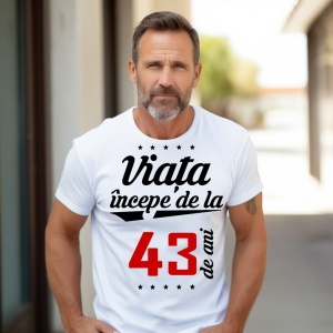 Bărbat matur cu barbă scurtă, purtând un tricou alb cu mesajul 'Viața începe de la 43 de ani', stând în exterior pe o alee însorită.