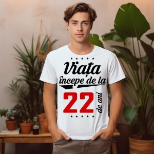 Tânăr purtând un tricou alb cu mesajul 'Viața începe de la 22 de ani', stând într-o cameră decorată cu plante.