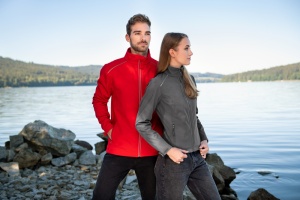Un bărbat și o femeie în jachete softshell, el în roșu și ea în gri, stau lângă o zonă stâncoasă la marginea unui lac, într-un peisaj natural cu dealuri în depărtare