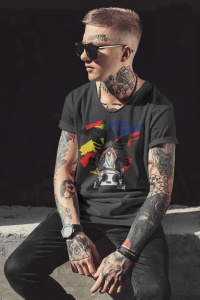 Bărbat tatuat, purtând ochelari de soare și un tricou negru cu un design urban, ce include un personaj cu un boombox și un fundal colorat abstract, pozând într-un cadru minimalist, luminat natural.