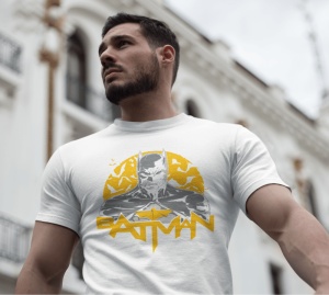 Bărbat purtând un tricou alb cu un design al personajului Batman, având un fundal galben cu lilieci și textul 'BATMAN' dedesubt. Este fotografiat într-un cadru urban, cu o clădire albă în fundal.