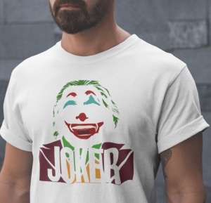 Prim-plan al unui bărbat purtând un tricou alb cu un design stilizat al personajului Joker, pe un fundal verde de iarbă. Designul include fața Jokerului zâmbind și textul 'JOKER' în partea de jos.