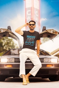 Bărbat purtând un tricou negru cu designul unei mașini sport și textul '911', așezat pe un DeLorean cu portierele deschise în sus, pozând într-un cadru retro cu un panou de benzinărie în fundal.