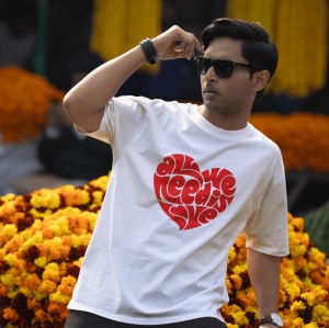 Bărbat purtând ochelari de soare și un tricou alb cu un design roșu în formă de inimă, conținând textul 'All we need is love', stând în fața unui fundal de flori portocalii și galbene.