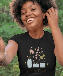 Tânără zâmbitoare purtând un tricou negru cu ilustrații de flori în vaze și mesajul 'you are about to bloom'. Ea are părul creț și poartă cerceri. Fundalul verde sugerează un mediu natural, iar fata pare să fie într-un cadru relaxat și vesel.