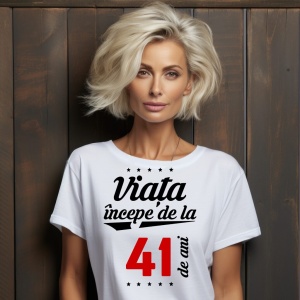 O femeie purtând un tricou alb cu textul  'Viața începe de la 41 de ani'. Fundal din lemn cu diferite nuanțe de maro și verde.