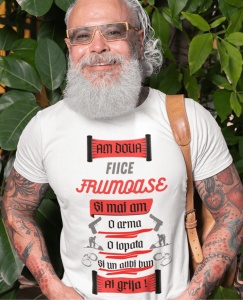 Bărbat zâmbitor, cu barbă albă și ochelari, purtând un tricou alb cu mesajul 'Am două fiice frumoase și mai am o armă, o lopată și un alibi bun. Ai grijă!', decorat cu ilustrații de arme, lopată și cătușe. Bărbatul are tatuaje pe brațe și o geantă pe umăr