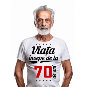 Bărbat în vârstă cu barbă albă, purtând un tricou alb cu mesajul 'Viața începe de la 70 de ani', pe un fundal alb simplu.