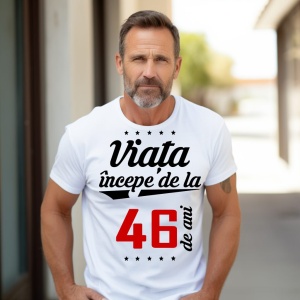 Bărbat matur cu barbă scurtă, purtând un tricou alb cu mesajul 'Viața începe de la 46 de ani', stând în exterior pe o alee însorită.