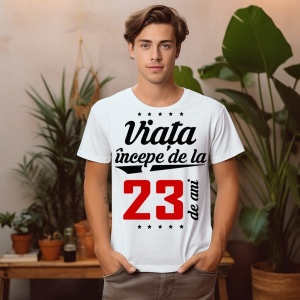 Tânăr purtând un tricou alb cu mesajul 'Viața începe de la 23 de ani', stând într-o cameră decorată cu plante.