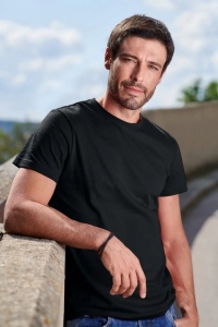 Bărbat sprijinit de un zid, purtând tricou negru, privind direct spre cameră în aer liber, pe fundalul unui cer senin