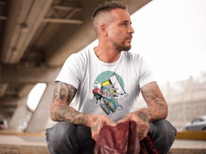 Bărbat tatuat purtând un tricou alb cu un design grafic colorat, stând pe vine sub o structură de pod modern, cu privirea îndreptată în lateral și ținând o geacă în mâini.