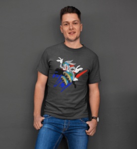Bărbat zâmbitor purtând un tricou negru cu un design colorat reprezentând un personaj animat într-o scenă dinamică, pozând relaxat într-un cadru interior cu un perete gri în fundal.