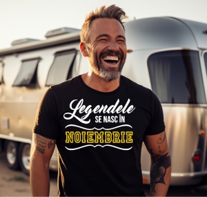 Un bărbat râzând, cu păr și barbă grizonate, purtând un tricou negru cu mesajul: „Legendele se nasc în Noiembrie”. În fundal se află o rulotă argintie, sugerând un cadru de vacanță sau aventură.