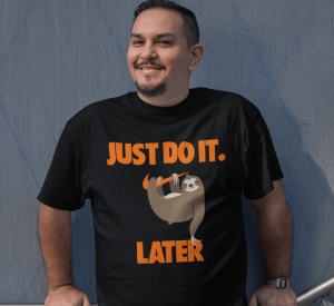 Un bărbat zâmbitor poartă un tricou negru cu un mesaj amuzant în portocaliu care spune 'JUST DO IT. LATER' și o ilustrație a unui leneș agățat de o ramură.
