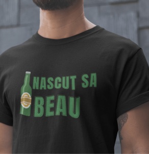 Tricou negru cu mesajul 'Născut să Beau', scris cu litere verzi, alături de o imagine stilizată a unei sticle de bere. Tricoul este purtat de un bărbat cu barbă, iar fundalul este neutru, cu un perete de culoare gri.