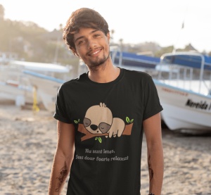 Un tânăr poartă un tricou negru cu o ilustrație drăguță a unui leneș care doarme pe o creangă, alături de textul 'Nu sunt leneș, Sunt doar foarte relaxat'. Tânărul zâmbește, iar fundalul arată o plajă cu bărci ancorate.