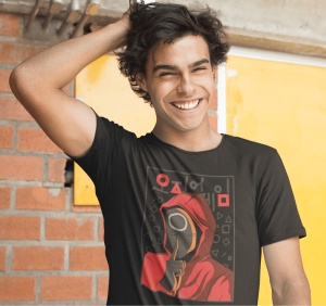 Un bărbat zâmbitor purtând un tricou negru cu un design inspirat din Squid Game, înfățișând un personaj cu mască roșie și simboluri geometrice, pozând în fața unui zid de cărămidă și a unei uși galbene.