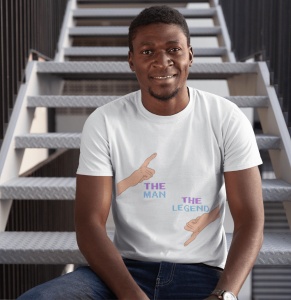 Un bărbat zâmbitor poartă un tricou alb cu un design amuzant ce include două mâini care arată cu degetul, una în sus cu textul 'The Man' și una în jos cu textul 'The Legend'. Bărbatul stă pe o scară metalică, fundalul fiind un cadru industrial.