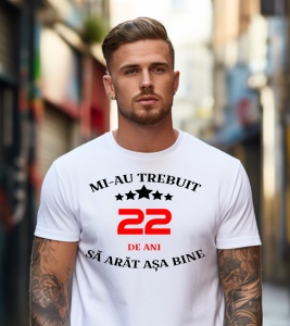 Un tânăr cu tatuaje pe braț, purtând un tricou alb cu mesajul în limba română: „Mi-au trebuit 22 de ani să arăt așa bine”, stă pe o stradă urbană.
